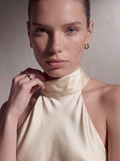 A close-up portrait of a model wearing a cream satin halter top and gold hoop earrings against a neutral beige background.